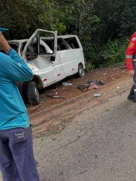 Van branca envolvida em acidente, estacionada à margem da estrada, com as portas abertas e algumas roupas e objetos espalhados ao redor. Duas pessoas, uma de camisa azul e outra com uniforme de emergência, estão próximas a ela.