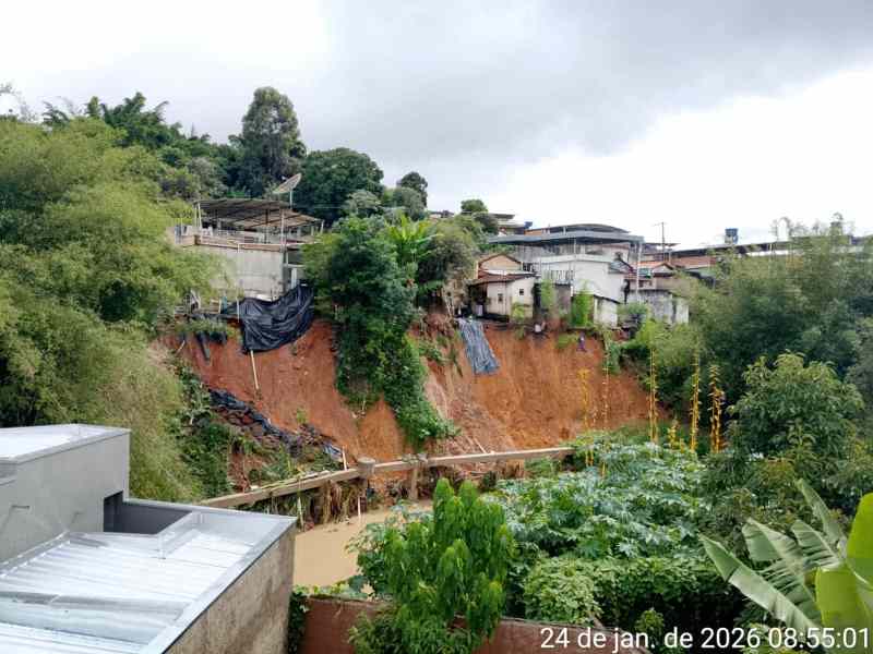 Vista de uma encosta com deslizamento de terra, casas ao fundo e vegetação em primeiro plano, em dia nublado.