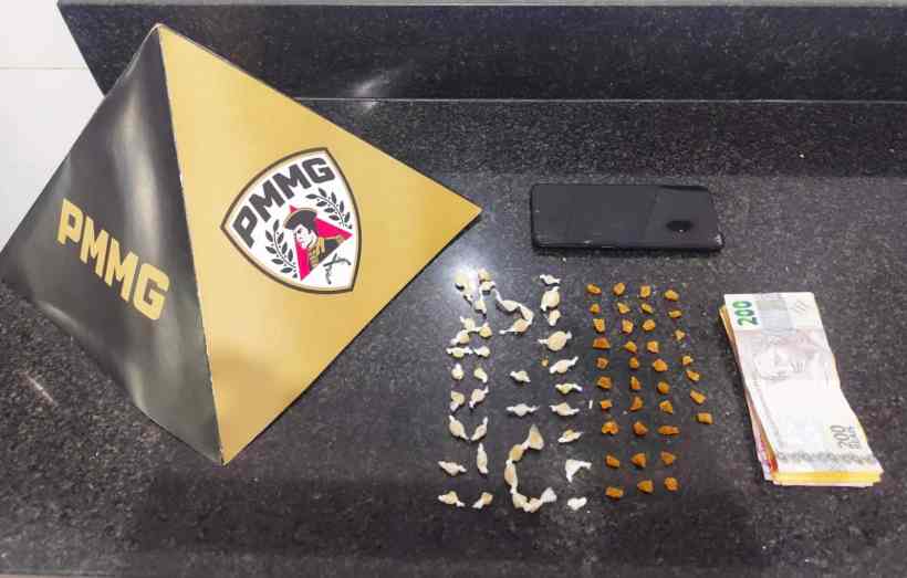 Uma bandeira da PMMG, um celular, várias porções de substâncias suspeitas e dinheiro empilhado sobre uma mesa.