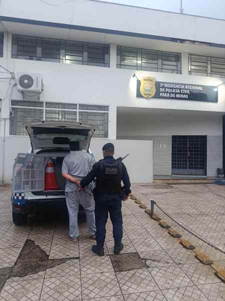 Um policial acompanha um homem em frente à 1ª Delegacia Regional de Polícia Civil de Minas Gerais, com um carro da polícia ao fundo.