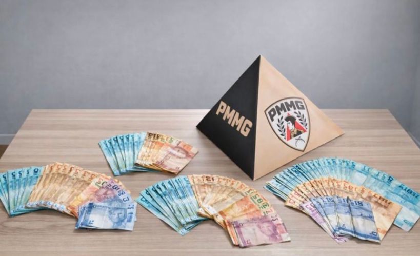 Notas de dinheiro em várias denominações dispostas ao redor de uma pirâmide com o logo da PMMG em uma mesa de madeira.