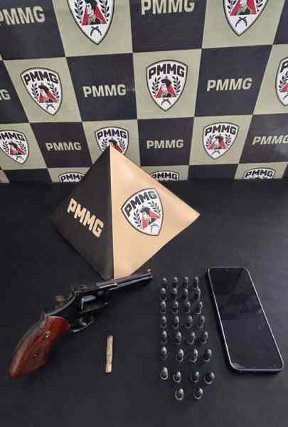Arma de fogo (revólver), celular, munições e objeto triangular com o logotipo PMMG sobre fundo com estande da PMMG.