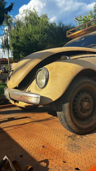Um fusca amarelo desgastado estacionado em uma superfície de metal laranja, com vegetação ao fundo.