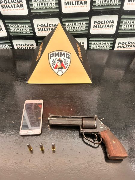 Uma arma de fogo tipo revólver ao lado de um celular e três cartuchos, com uma placa da Polícia Militar ao fundo.
