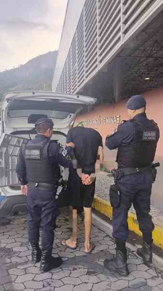 Três policiais em pé em frente a um veículo, um deles algemando um homem com as mãos atrás das costas.