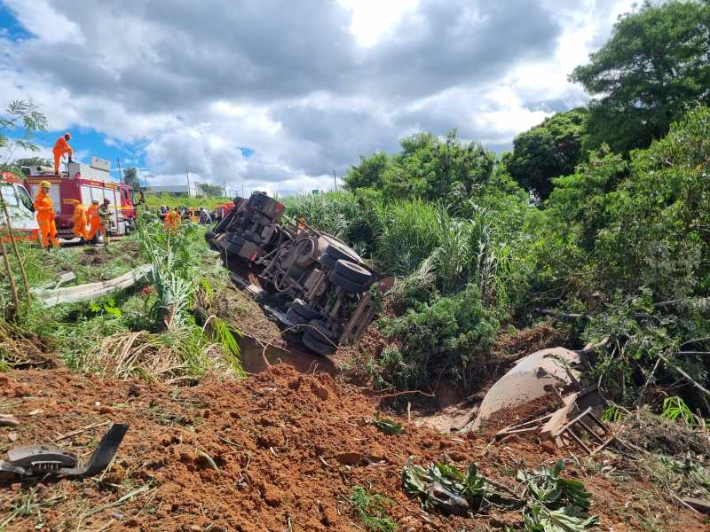 Caminhão tombado em um acostamento, com equipes de emergência trabalhando ao fundo em um dia ensolarado.
