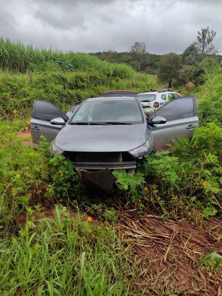Carro acidentado em uma área rural, cercado por vegetação alta, com as portas abertas.