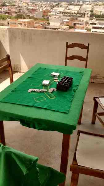 A green tablecloth-covered table on a balcony with scattered game pieces and a box, overlooking a city landscape.