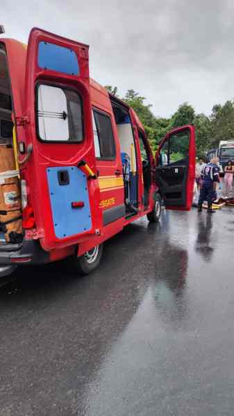 Ambulância vermelha com as portas abertas, estacionada em uma estrada molhada, enquanto profissionais de emergência se preparam para atender uma ocorrência.