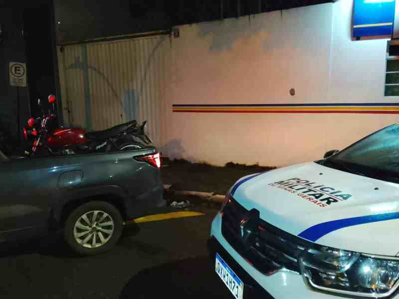 Um carro estacionado em uma rua à noite, com uma motocicleta sobre o banco traseiro. Um veículo da Polícia Militar está estacionado próximo ao local.