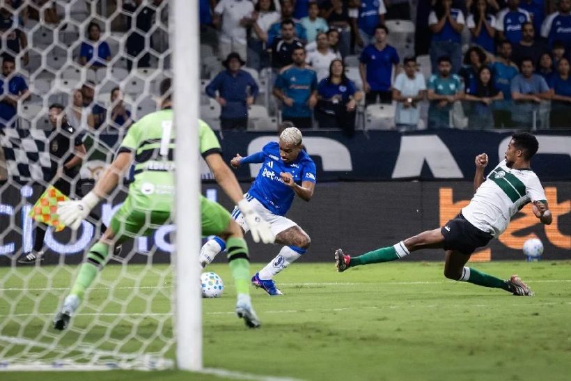 A soccer match scene featuring a player in blue attempting to evade a sliding tackle from an opponent in white and green, while the goalkeeper in green watches from the goal.