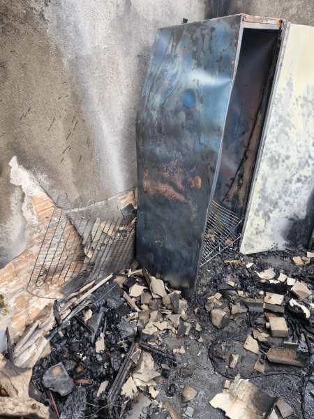 Cenário de um local com danos por incêndio, mostrando um refrigerador queimado e destroços ao redor, incluindo materiais carbonizados e um painel de parede danificado.