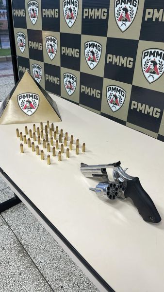 Uma mesa com um revólver, cartuchos de munição dispostos em padrão e uma pirâmide metálica ao fundo, em meio a um ambiente institucional.