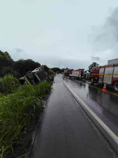 Caminhão tombado na rodovia, com viaturas de emergência ao fundo, em um dia chuvoso.