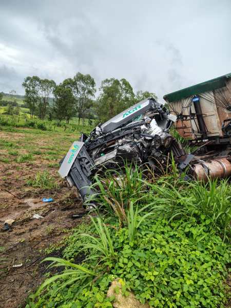 Caminhão acidentado deitado em um terreno com vegetação, rodeado por árvores e céu nublado.