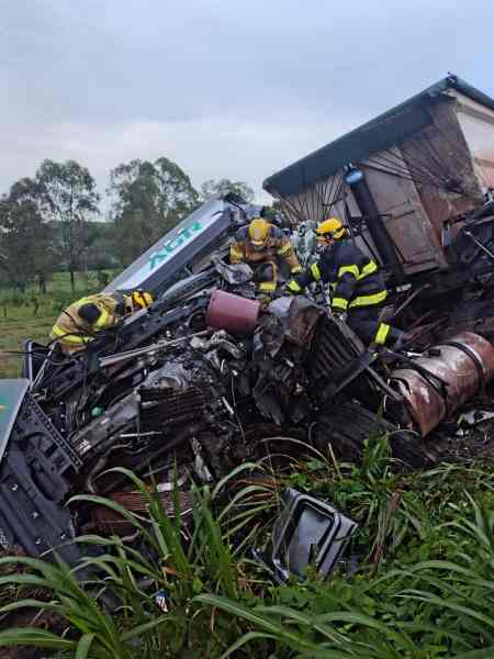 Bombeiros trabalhando em um caminhão tombado em uma área rural, com vegetação ao redor.