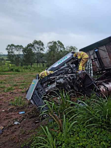 Caminhão acidentado com parte da carroceria tombada em um campo, enquanto bombeiros realizam o resgate e a contenção da situação.