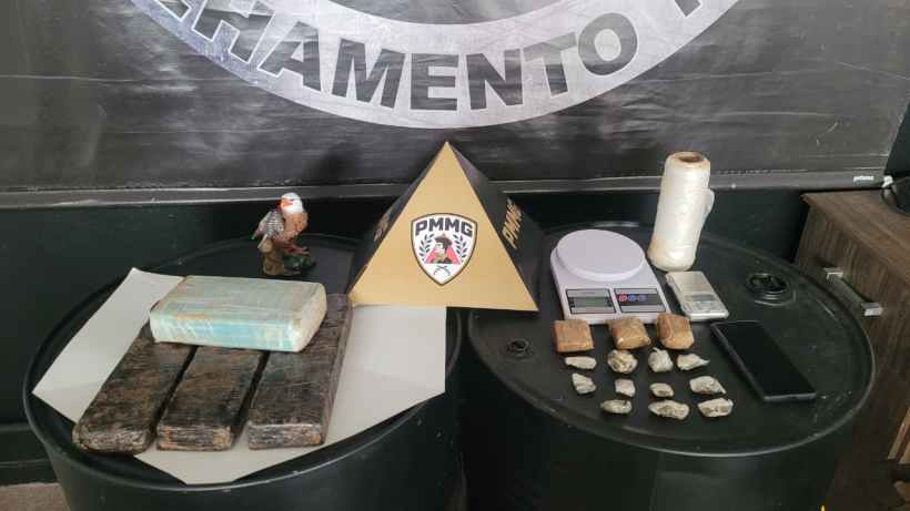 Mesa com objetos relacionados a investigação, incluindo um bloco de madeira, balança, rolo de linha e pedras, ao lado de um símbolo da PMMG.