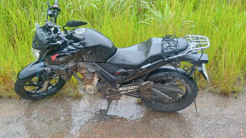 Motocicleta preta com sujeira e molhada, estacionada em uma estrada de terra ao lado de capim alto.