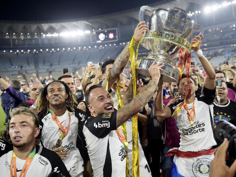 A group of soccer players celebrating with a trophy in a stadium, surrounded by fans and confetti.