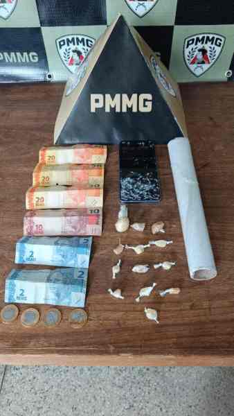 Imagem de materiais apreendidos, incluindo dinheiro em várias notas, uma faca, um celular e embalagens com substâncias suspeitas, dispostos sobre uma mesa e com um fundo de parede de polícia.