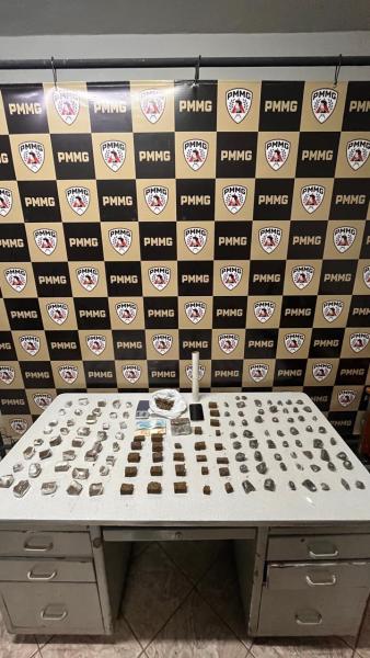 Mesa com diversos objetos, incluindo pacotes e itens organizados, em frente a um painel com logotipos da PMMG.