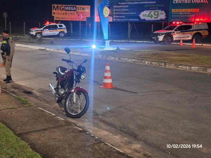 Imagem noturna de uma rua com uma moto vermelha estacionada à frente. No fundo, há um veículo da polícia e uma placa iluminada. Um policial está presente na cena.