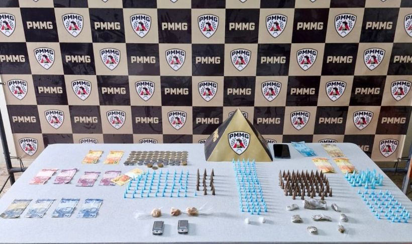 Mesas com dinheiro, drogas e celulares dispostos, com uma parede de fundo com logotipos da PMMG.