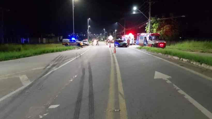 Cena noturna de uma estrada com marcas de pneus, onde veículos de emergência e polícia estão parados, com luzes piscando.