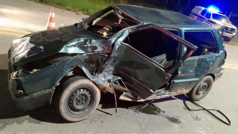 Carro verde com a parte frontal danificada e porta aberta, abandonado em uma estrada à noite, com cones de sinalização e viatura ao fundo.