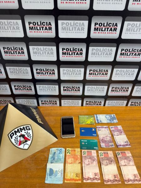 Foto de uma mesa com dinheiro, cartões de crédito e um celular, em frente a um fundo com logos da Polícia Militar de Minas Gerais.