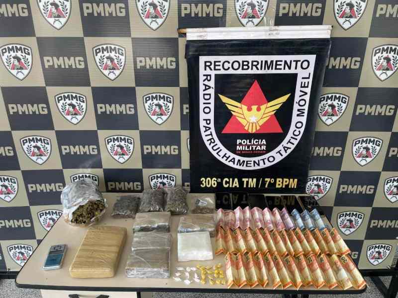 Mesa com drogas apreendidas, incluindo pacotes de maconha, substâncias em pó e comprimidos, com um banner da Polícia Militar ao fundo.