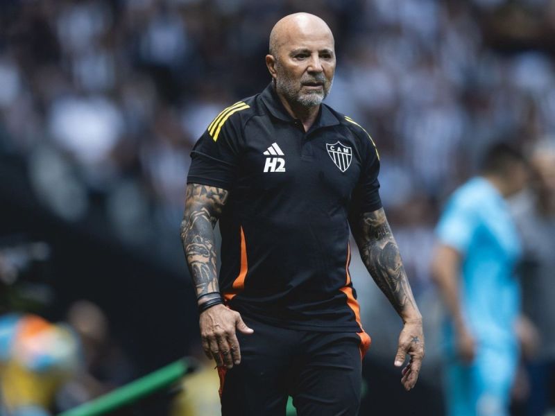 A football coach with tattoos wearing a black team tracksuit with orange accents, walking on the pitch during a match.