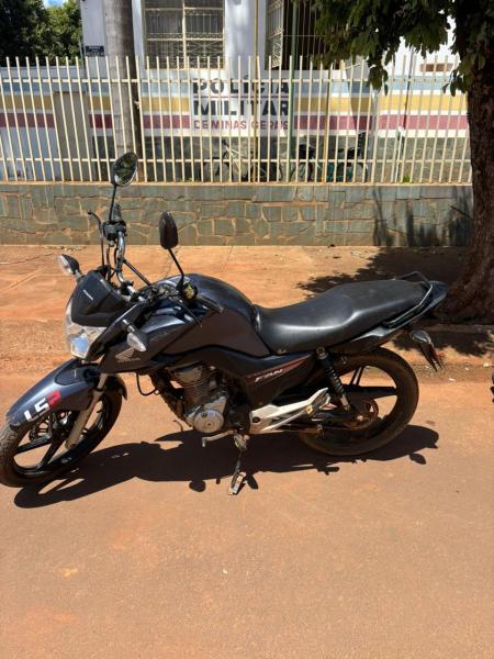 Uma motocicleta preta estacionada na calçada, com um fundo que mostra um prédio da polícia.