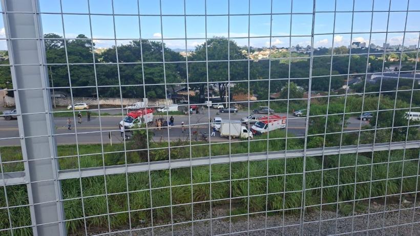 Vista de um acidente de trânsito com ambulâncias e equipes de atendimento em uma estrada, vista através de uma grade de proteção.