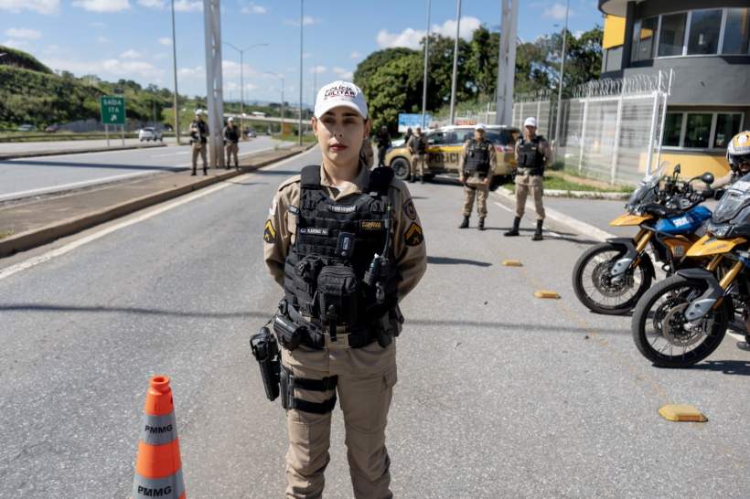 Policial feminina em uniforme, com colete tático, posando na estrada com veículos de polícia ao fundo.