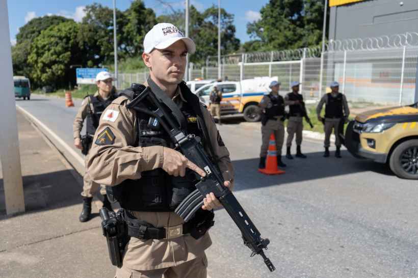 Policial em uniforme tático segurando uma arma, com outros oficiais ao fundo e veículos de polícia estacionados.