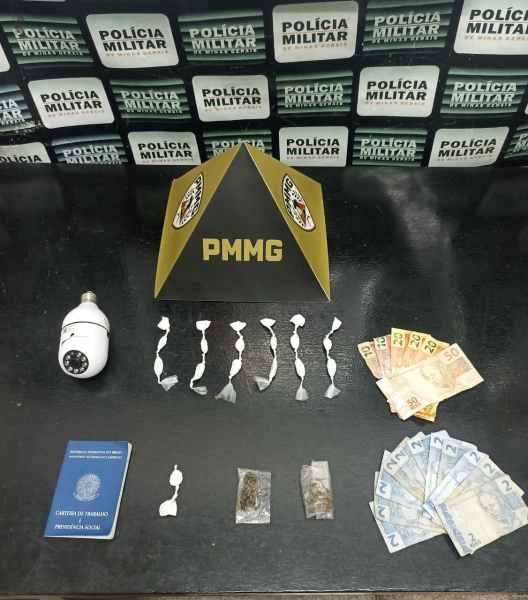 Material apreendido em operação policial, incluindo drogas embaladas, dinheiro, um passaporte e um dispositivo de segurança. Em fundo com logo da Polícia Militar.