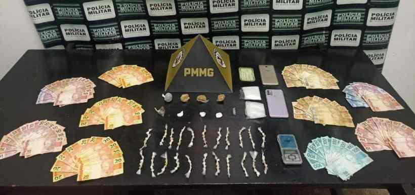 Dinheiro em várias denominações, objetos suspeitos e telefones móveis dispostos em uma mesa com fundo da Polícia Militar.