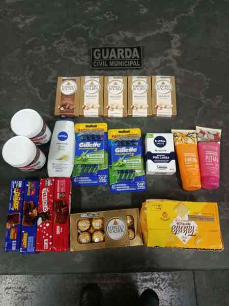 Imagem mostrando produtos diversos, incluindo chocolates, itens de higiene pessoal e cremes, dispostos sobre uma superfície. No fundo, há uma placa escrita 'GUARDA CIVIL MUNICIPAL'.