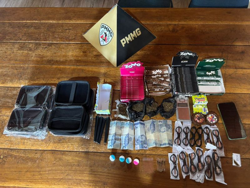 Mesa de madeira com itens diversos: um triângulo dourado com a inscrição PMMG, caixas de folhas de papel, sacos com substâncias escuras, dinheiro em notas, balanças, e outros acessórios relacionados.