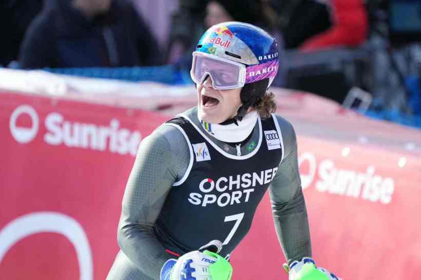 A skier wearing a helmet and goggles celebrates at a skiing event, with a backdrop featuring sponsor logos.