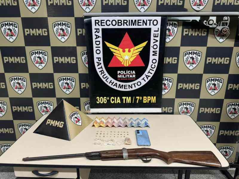 Imagem de uma mesa com uma espingarda, um celular, diversos envelopes coloridos e um display com informações da Polícia Militar.