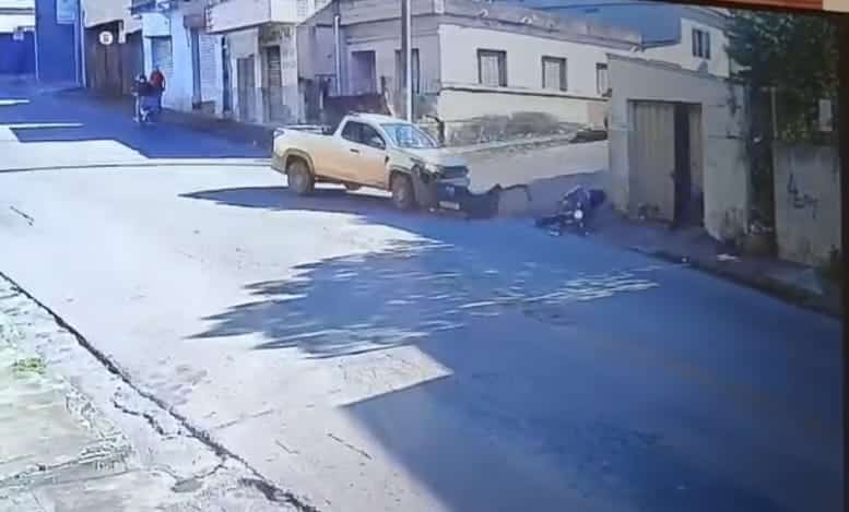 Uma cena de rua mostrando um carro amarelo estacionado em uma calçada, com uma pessoa próxima a ele e algumas casas ao fundo.