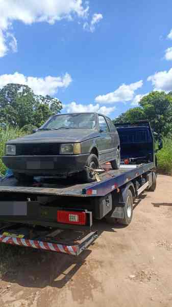 Um carro preto sendo transportado em um reboque em uma estrada de terra, com árvores e céu azul ao fundo.