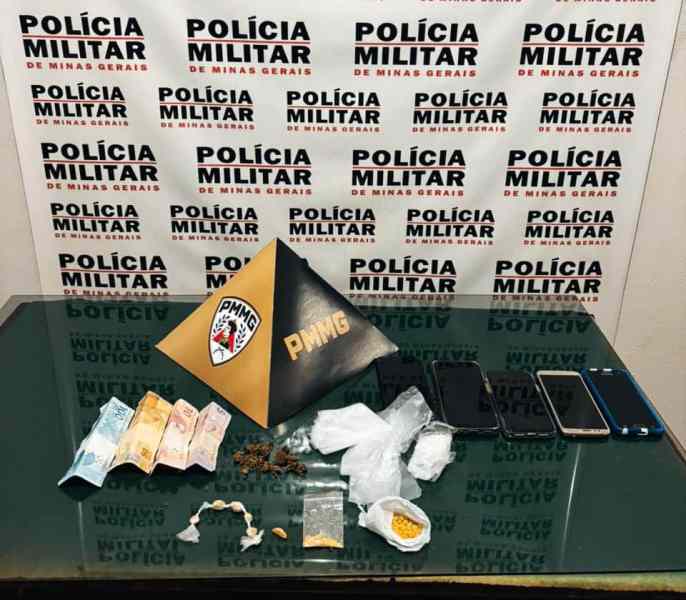 Imagem de material apreendido pela Polícia Militar de Minas Gerais, incluindo drogas, dinheiro e celulares, exibidos sobre uma superfície vidro com fundo de banner da polícia.