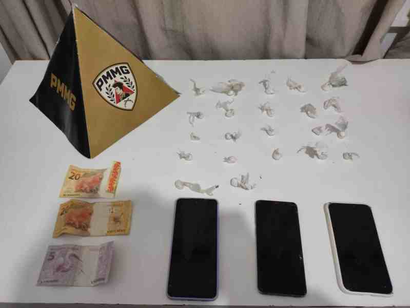 Pyramid com o logotipo da PMMG, três smartphones pretos e um branco, dinheiro em notas de R$ 5 e R$ 20, e pequenos papéis em uma mesa.