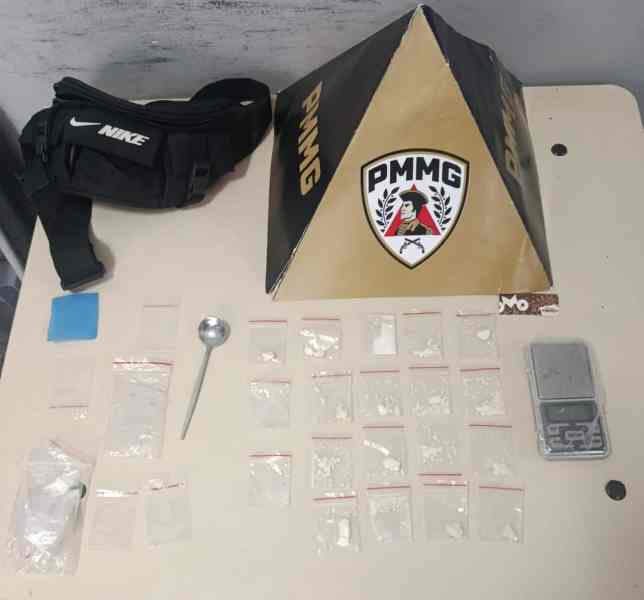 Mesa com uma bolsa preta, um objeto em forma de pirâmide com o logotipo PMMG, uma colher e várias sacolas plásticas contendo substâncias brancas.