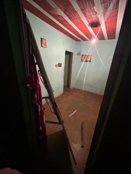 Um interior de quarto com paredes verdes e teto vermelho, apresentando uma lâmpada acesa que projeta luz sobre o chão de terra. Duas portas levam a outros cômodos e há algumas imagens penduradas nas paredes.