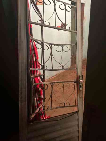 Uma porta de metal com grade aberta, mostrando um interior simples, com chão de terra e paredes sem revestimento. Atrás da porta, há um pano vermelho pendurado.